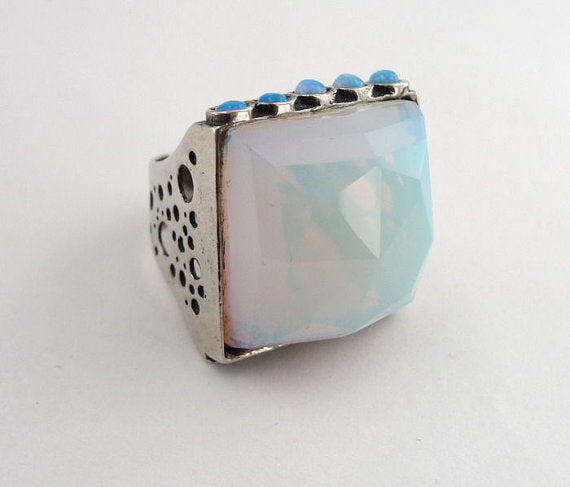 Bold sterling silver ring featuring a large, faceted 20mm square Opalite gemstone as the centerpiece, accented by five small blue opal stones along the top edge. The ring’s substantial design includes a textured silver band with openwork detailing, creating a unique and artistic look.