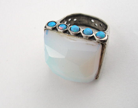 Bold sterling silver ring featuring a large, faceted 20mm square Opalite gemstone as the centerpiece, accented by five small blue opal stones along the top edge. The ring’s substantial design includes a textured silver band with openwork detailing, creating a unique and artistic look.