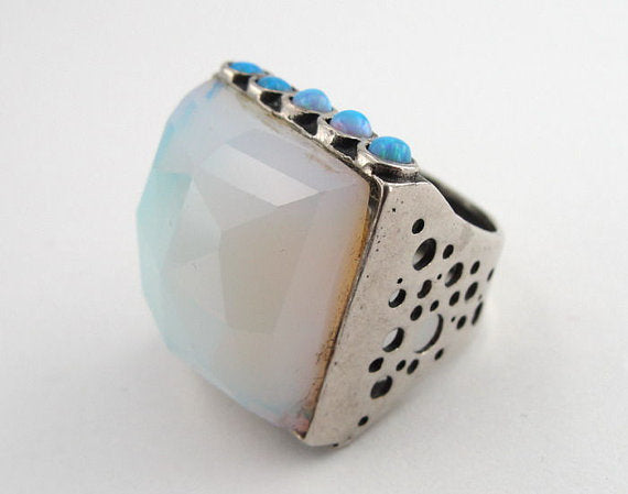 Bold sterling silver ring featuring a large, faceted 20mm square Opalite gemstone as the centerpiece, accented by five small blue opal stones along the top edge. The ring’s substantial design includes a textured silver band with openwork detailing, creating a unique and artistic look.