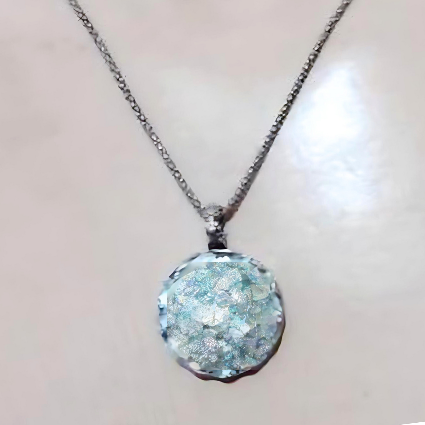 Handcrafted sterling silver pendant featuring a round Roman glass centerpiece in shades of light blue and silver. The unique, textured chain complements the pendant, creating a distinctive, artisanal look. Shown held in hand to display its intricate details and delicate craftsmanship