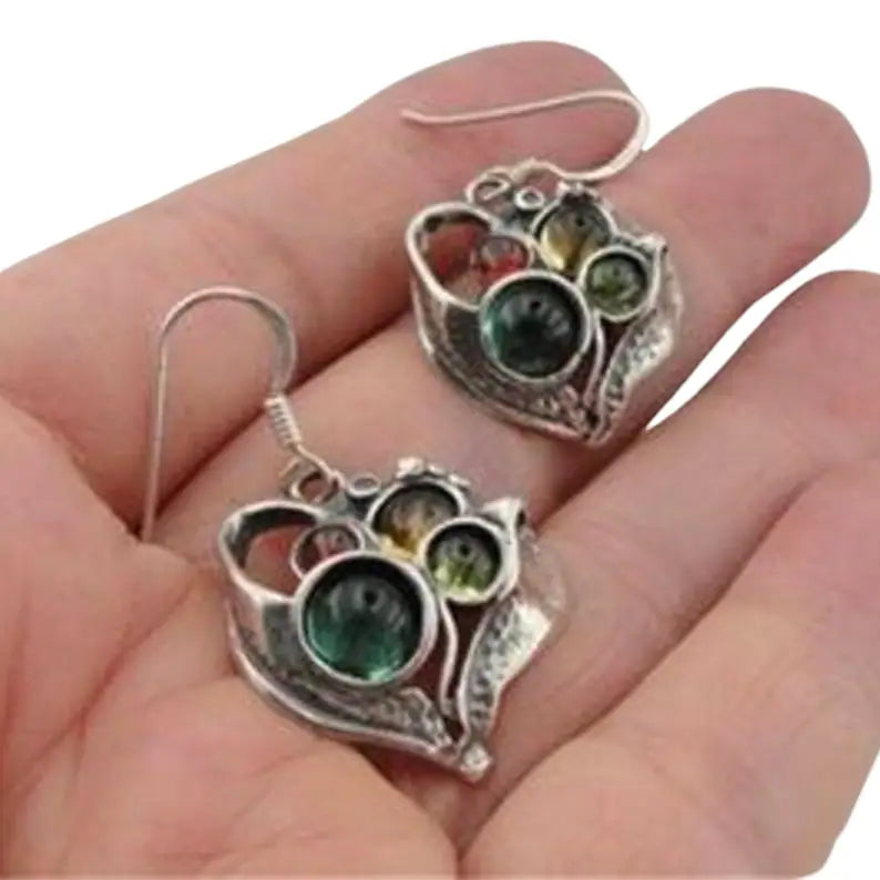 Close-up view of elegant, handcrafted heart-shaped silver earrings, featuring a unique, intricate design with multicolored glass beads in shades of emerald green, amber, and golden yellow. The earrings showcase a vintage-inspired aesthetic with a delicate arrangement of glass stones, adding a touch of sophistication and artistic charm