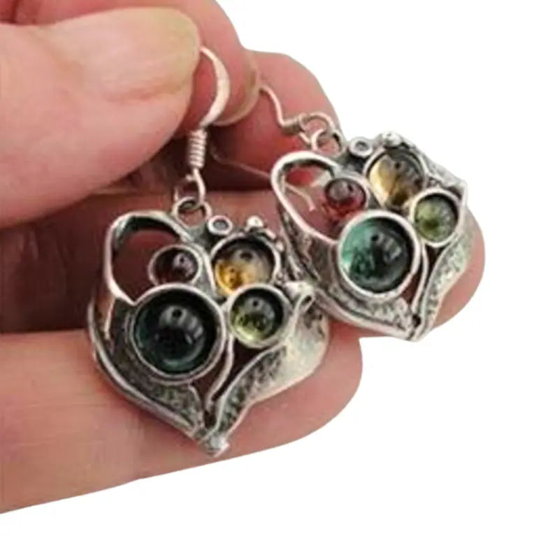 Close-up view of elegant, handcrafted heart-shaped silver earrings, featuring a unique, intricate design with multicolored glass beads in shades of emerald green, amber, and golden yellow. The earrings showcase a vintage-inspired aesthetic with a delicate arrangement of glass stones, adding a touch of sophistication and artistic charm