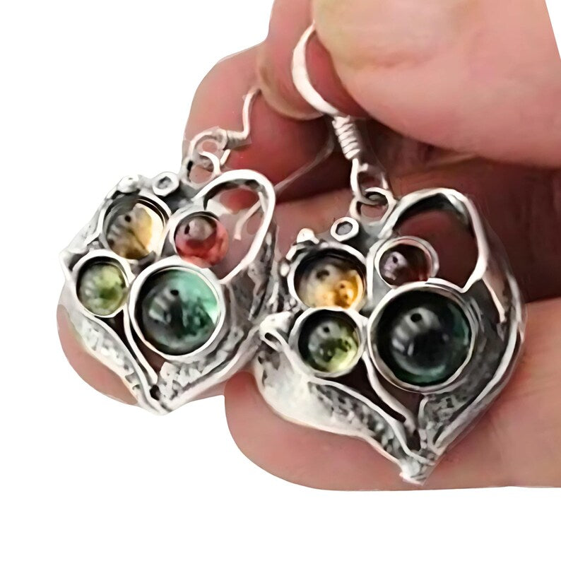 Close-up view of elegant, handcrafted heart-shaped silver earrings, featuring a unique, intricate design with multicolored glass beads in shades of emerald green, amber, and golden yellow. The earrings showcase a vintage-inspired aesthetic with a delicate arrangement of glass stones, adding a touch of sophistication and artistic charm