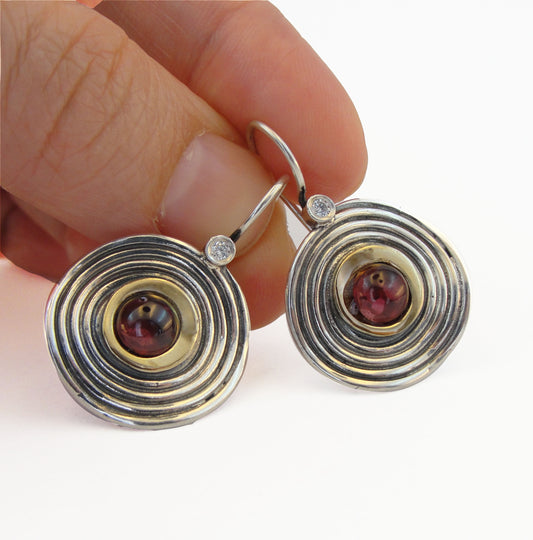 sterling silver earrings with concentric circle design featuring 9K yellow gold accents and round garnet gemstones at the center. Each earring is adorned with a small clear zircon stone on the hook, creating an elegant and modern look. Perfect for adding a touch of sophistication to any outfit, these artisan-crafted earrings showcase quality craftsmanship and unique design.